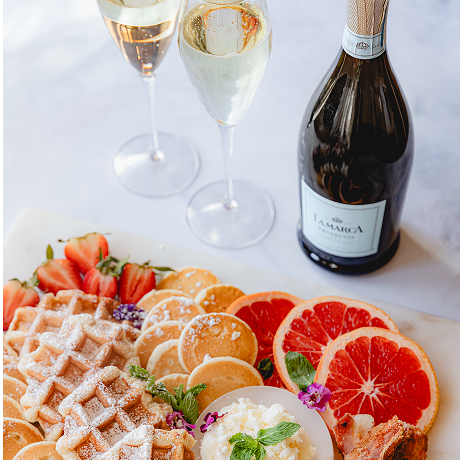 A gourmet brunch spread on a marble table featuring waffles, mini pancakes, sliced grapefruit, and strawberries, accompanied by two filled champagne flutes and a bottle of La Marca Prosecco.