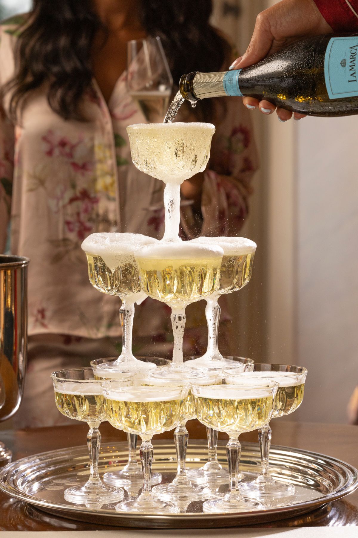 A close-up shot of a champagne tower made of stacked coupe glasses as La Marca Prosecco is being poured from the top, with bubbles and effervescence overflowing against a soft, bright background.