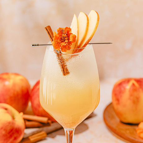 A sparkling light-orange cocktail in a wine glass garnished with three fresh apple slices, a cinnamon stick, and a single orange marigold flower, with whole apples in the soft-focus background.