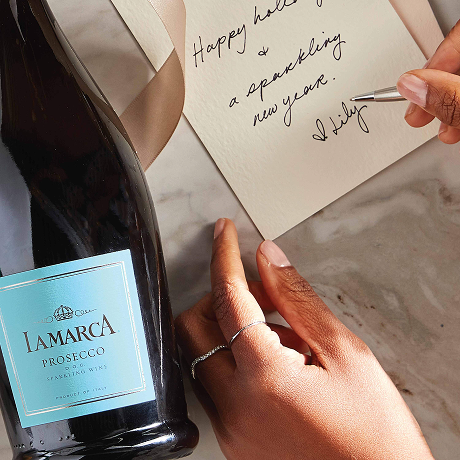 Top-down view of a hand writing a holiday card that reads 'Happy holidays and a sparkling new year' next to a bottle of La Marca Prosecco on a marble surface.