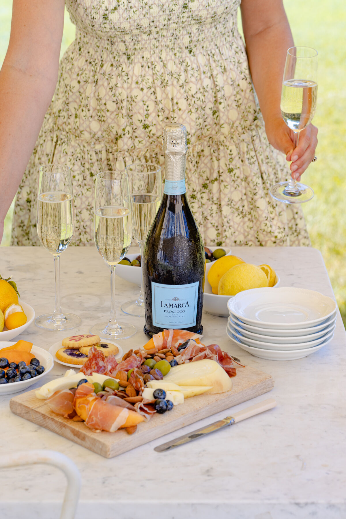 La Marca Prosecco bottle on a table with filled champagne flutes and a charcuterie board with cheese, cured meats, olives, and fruit, with a person holding a glass in the background