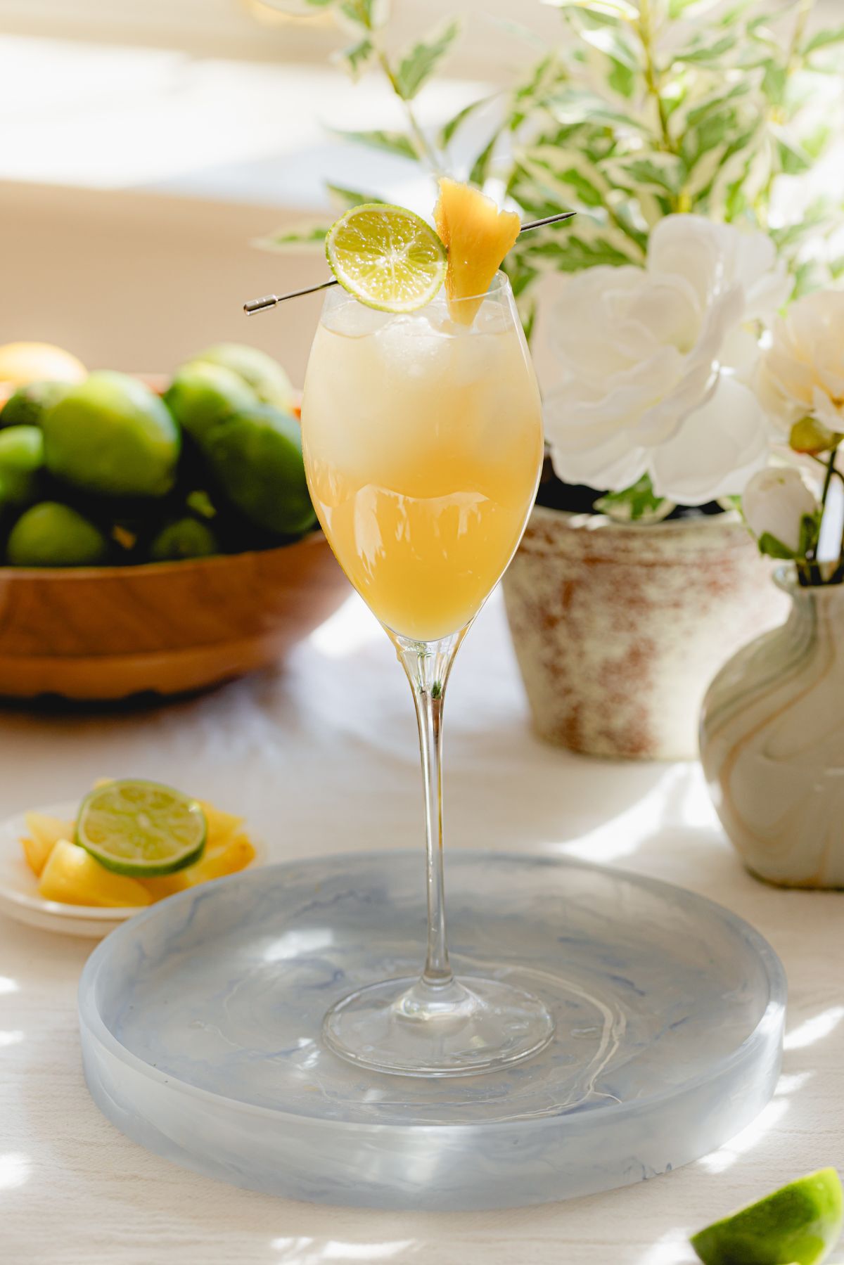 Golden Candied Sapphire Prosecco cocktail garnished with lime and pineapple, served in a flute glass alongside fresh citrus and floral accents. sapphire