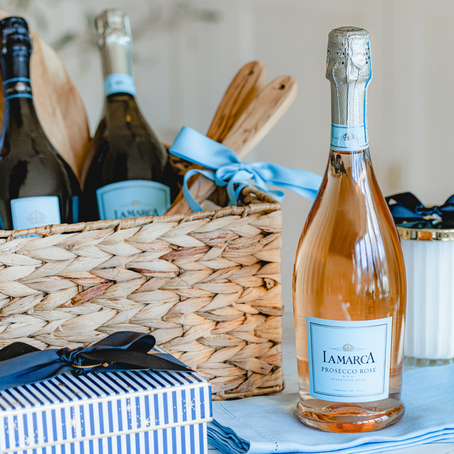 A bottle of La Marca Prosecco Ros&eacute; stands prominently in the foreground, with a curated wicker gift basket in the soft-focus background containing bottles of La Marca Prosecco DOC and Luminore, accented by white flowers and a light blue ribbon.