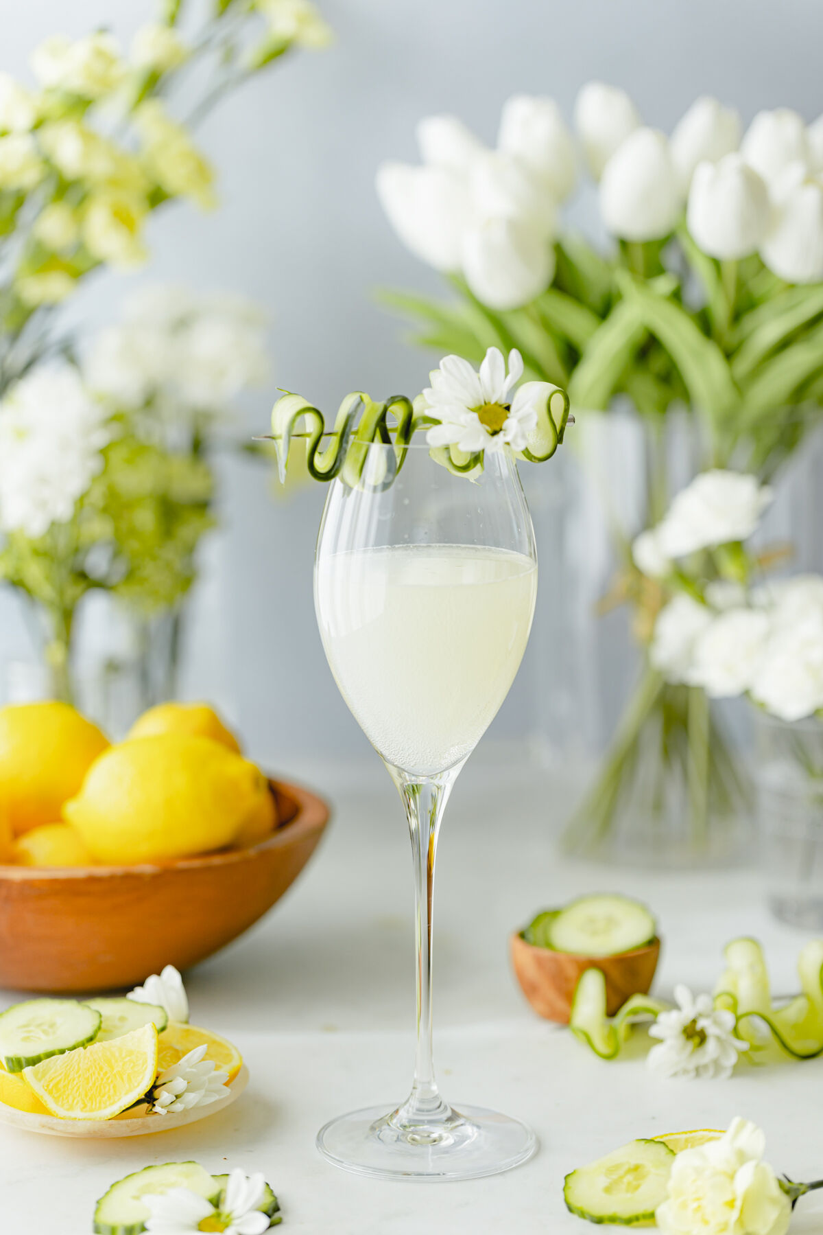 Queen Bee Prosecco cocktail garnished with cucumber ribbon and white flower, surrounded by lemons, cucumber slices, and fresh floral arrangements.