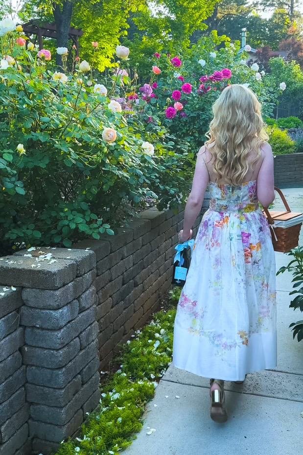 Woman in a floral dress carrying a bottle of La Marca Prosecco and picnic basket while walking through a blooming rose garden.