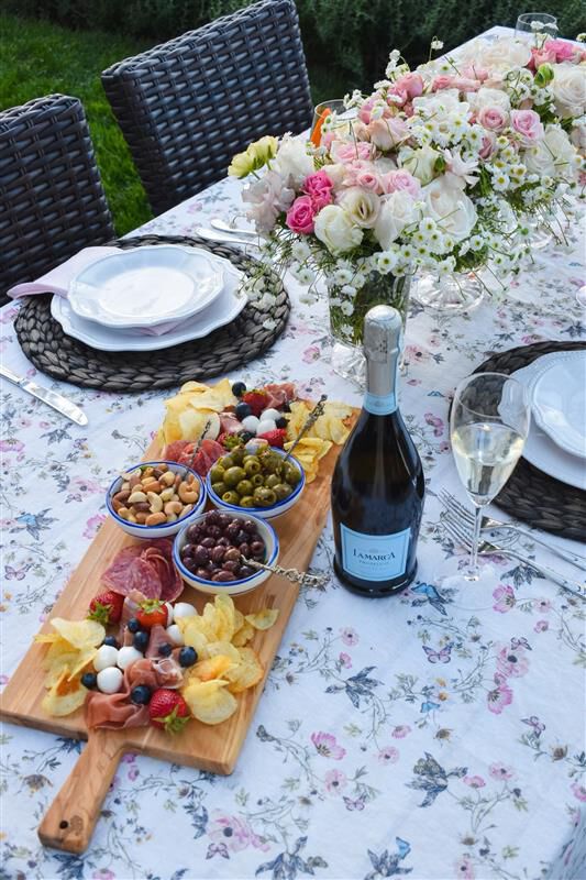 Charcuterie board with fruit, olives, cheeses, and cured meats served alongside a bottle and glass of La Marca Prosecco on a floral-covered outdoor table.